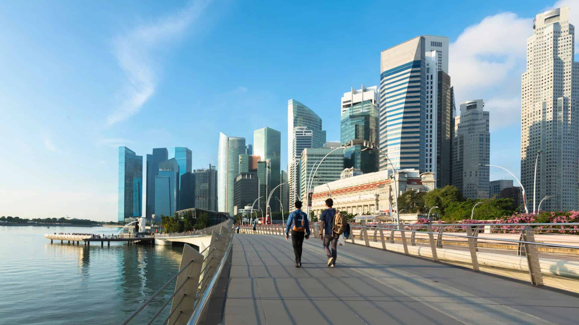 Two people walk on a bridge toward a modern city skyline with tall glass buildings by the water.