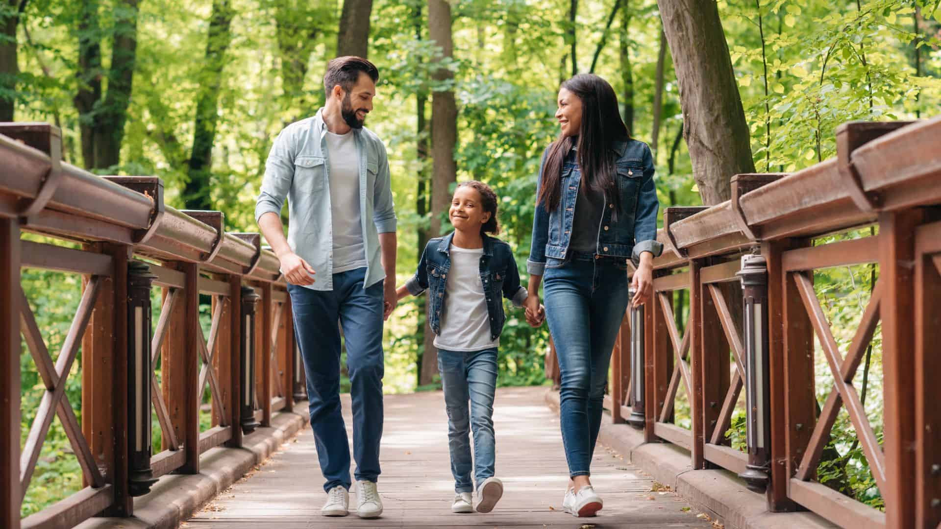 A family of three walks together on a wooden bridge in a lush green forest, smiling and holding hands.