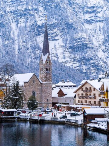 Snow-covered village with colorful buildings and a church by a lake, surrounded by snowy mountains in winter.
