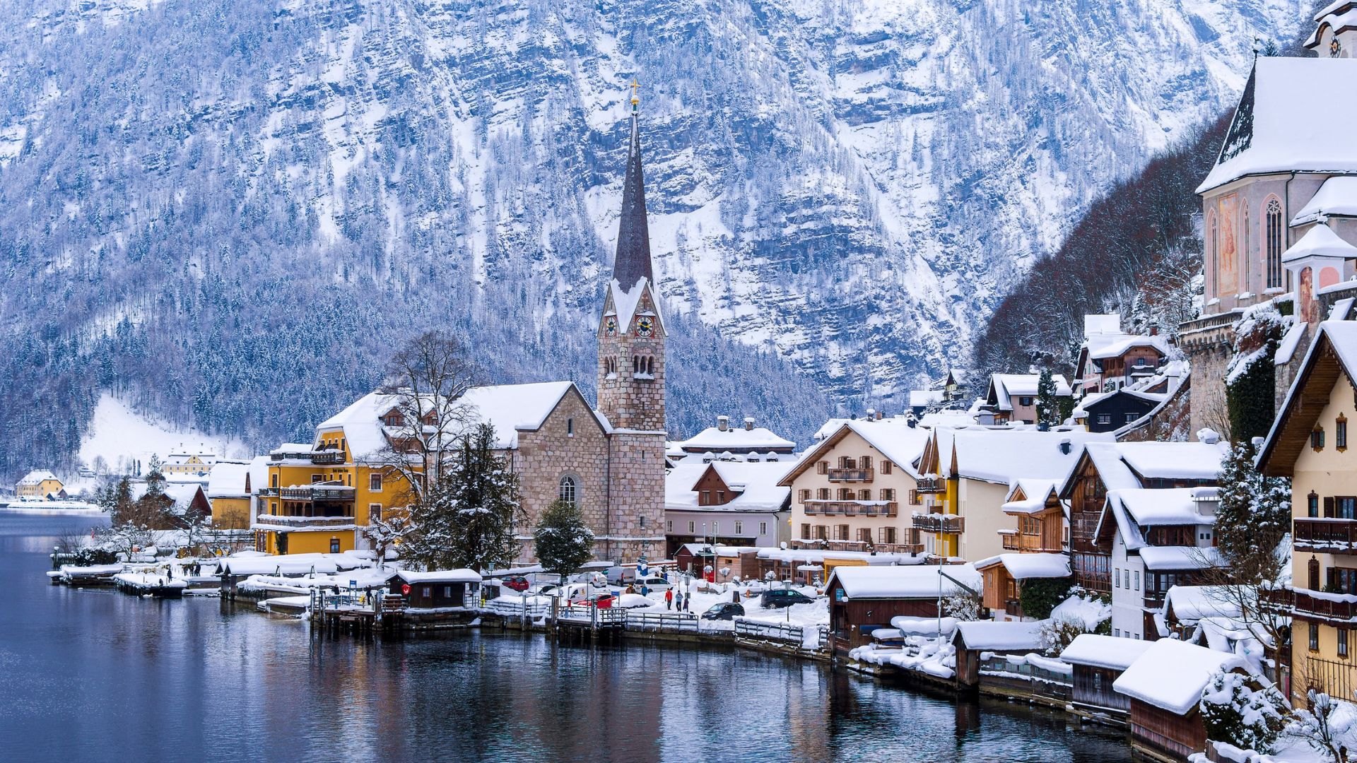 Snow-covered village with colorful buildings and a church by a lake, surrounded by snowy mountains in winter.