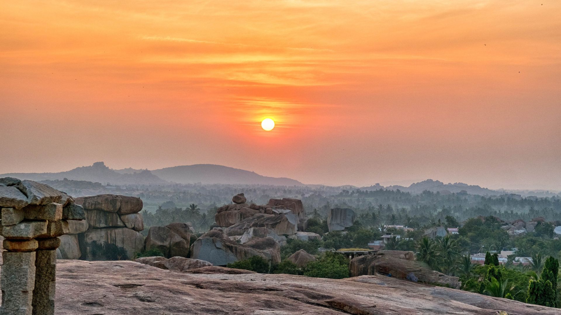 Sunset over rocky hills and greenery, with the sun low in an orange sky above a distant village.