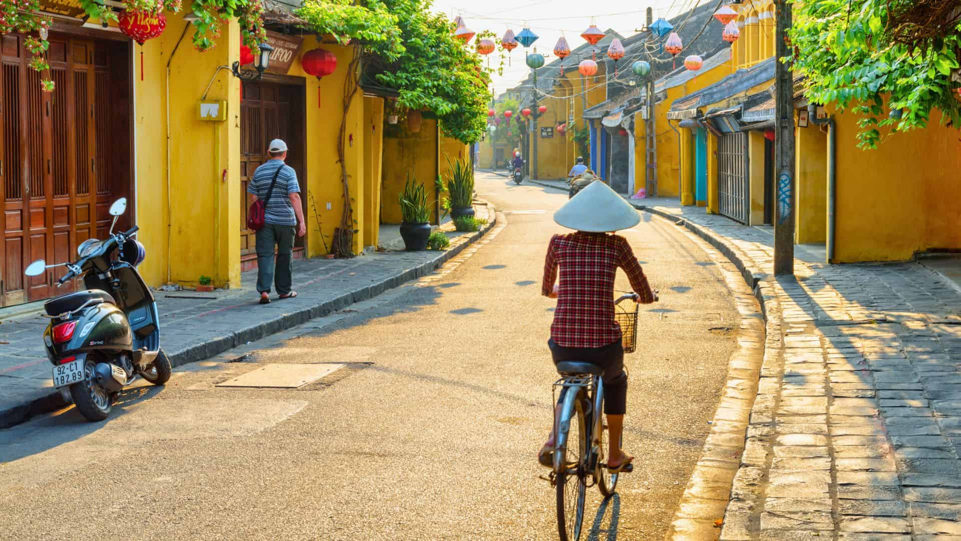 Person in conical hat rides bicycle down a sunny, lantern-lined street with yellow buildings in Hoi An, Vietnam.