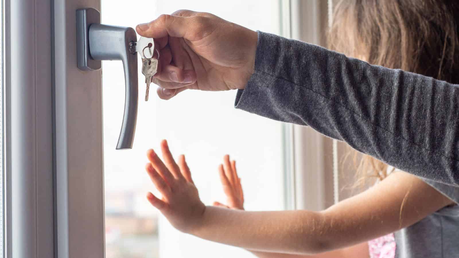 An adult locking a window with a key while a child stands nearby with hands on the glass.