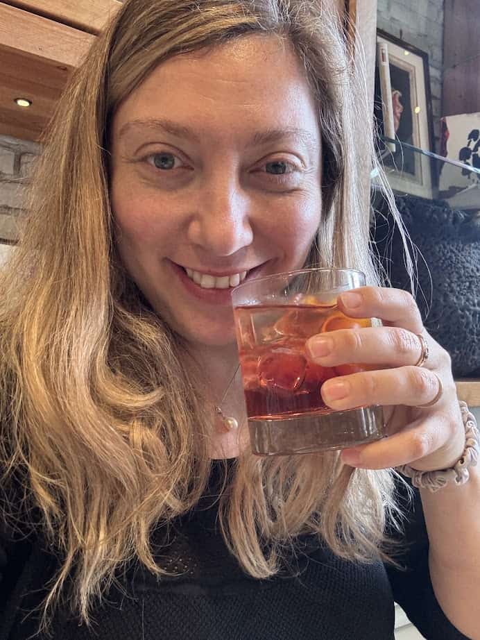 Smiling woman with long blonde hair holding a cocktail with ice in a glass, enjoying the vibrant atmosphere near Barcelona markets.
