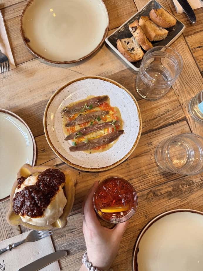 A hand holds a red drink next to fresh food appetizers, bread, and empty plates on a rustic wooden table—a perfect scene for those exploring where to eat at Barcelona markets.