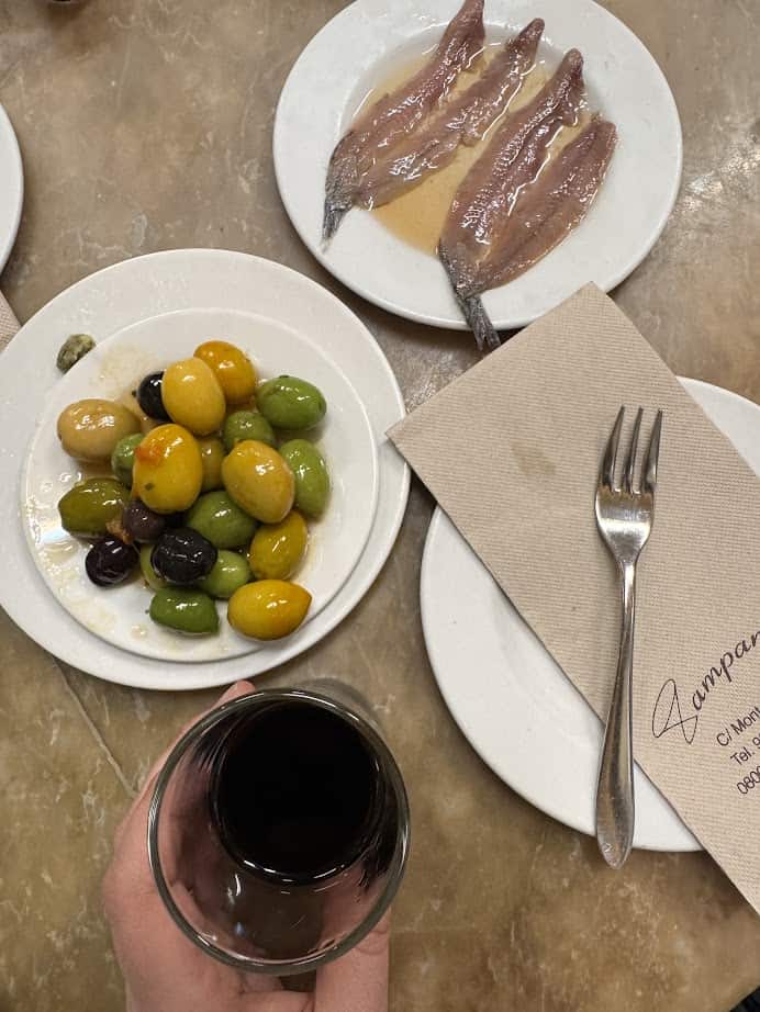 A hand holding a glass of red wine next to plates of olives and anchovies on a marble table, capturing the spirit of fresh food found in Barcelona markets.