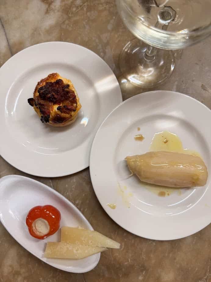 Three plates with tapas—stuffed pepper, cheese slices, baked pastry, and an endive with sauce—plus a wine glass offer a taste of fresh food you’ll find at Barcelona markets, perfect for anyone wondering where to eat in the city.