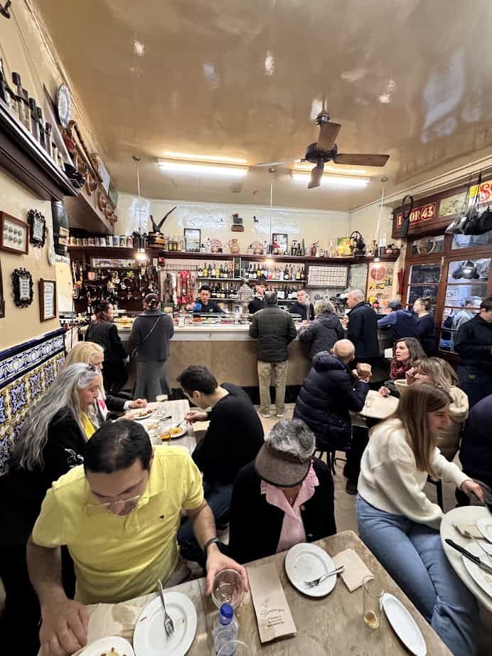 Busy tapas bar with people eating and chatting at tables and the counter, warm lighting, cozy atmosphere&mdash;perfect for those seeking where to eat fresh food like you'd find in Barcelona markets.