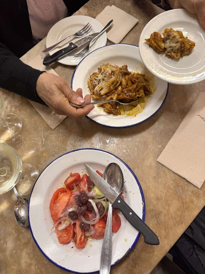 Two people serve baked pasta and a tomato salad with olives at a restaurant table, highlighting the fresh food inspired by Barcelona markets.