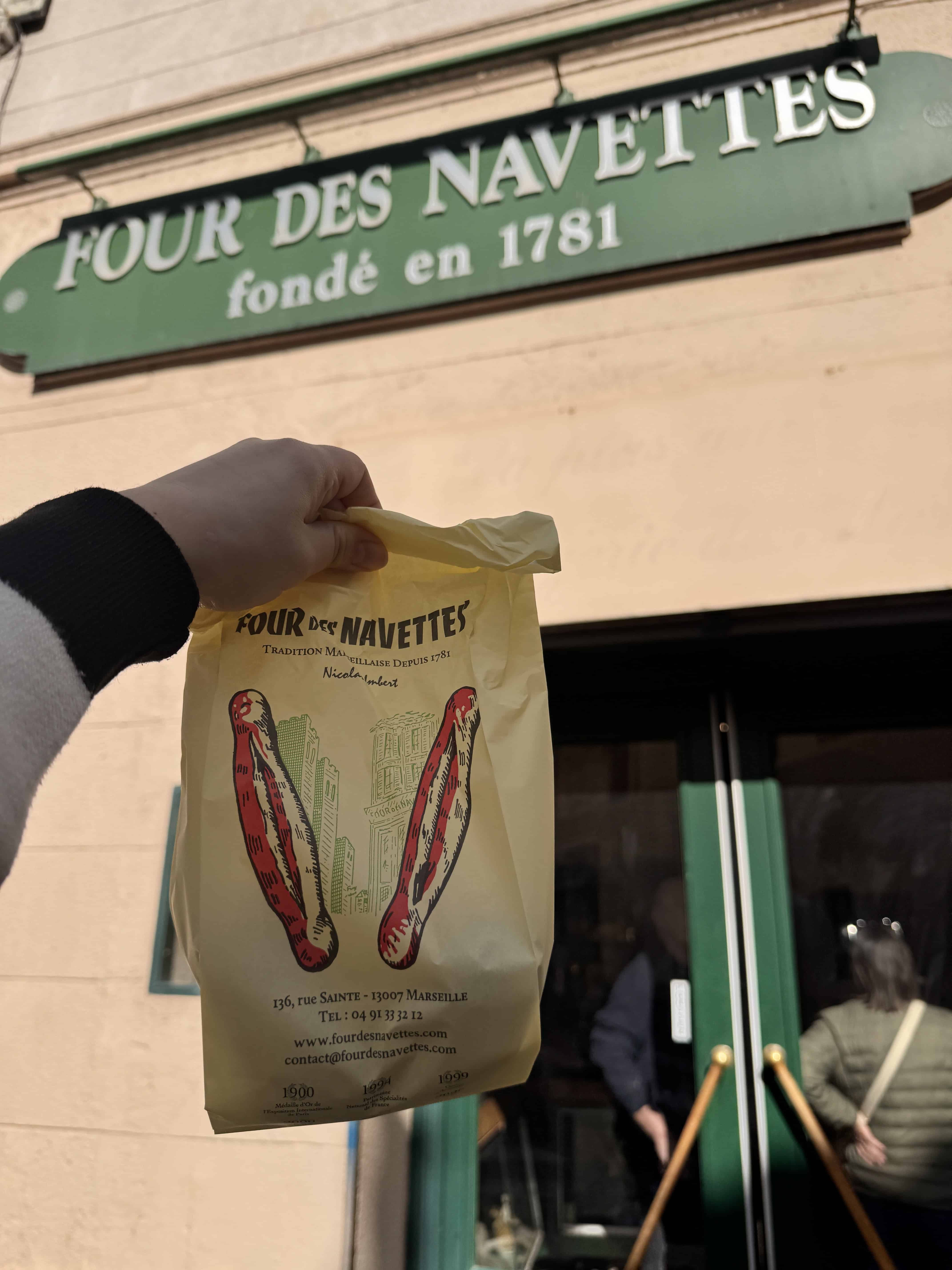 A hand holds a yellow pastry bag in front of Four des Navettes bakery—a must-stop on any Marseille itinerary in France.