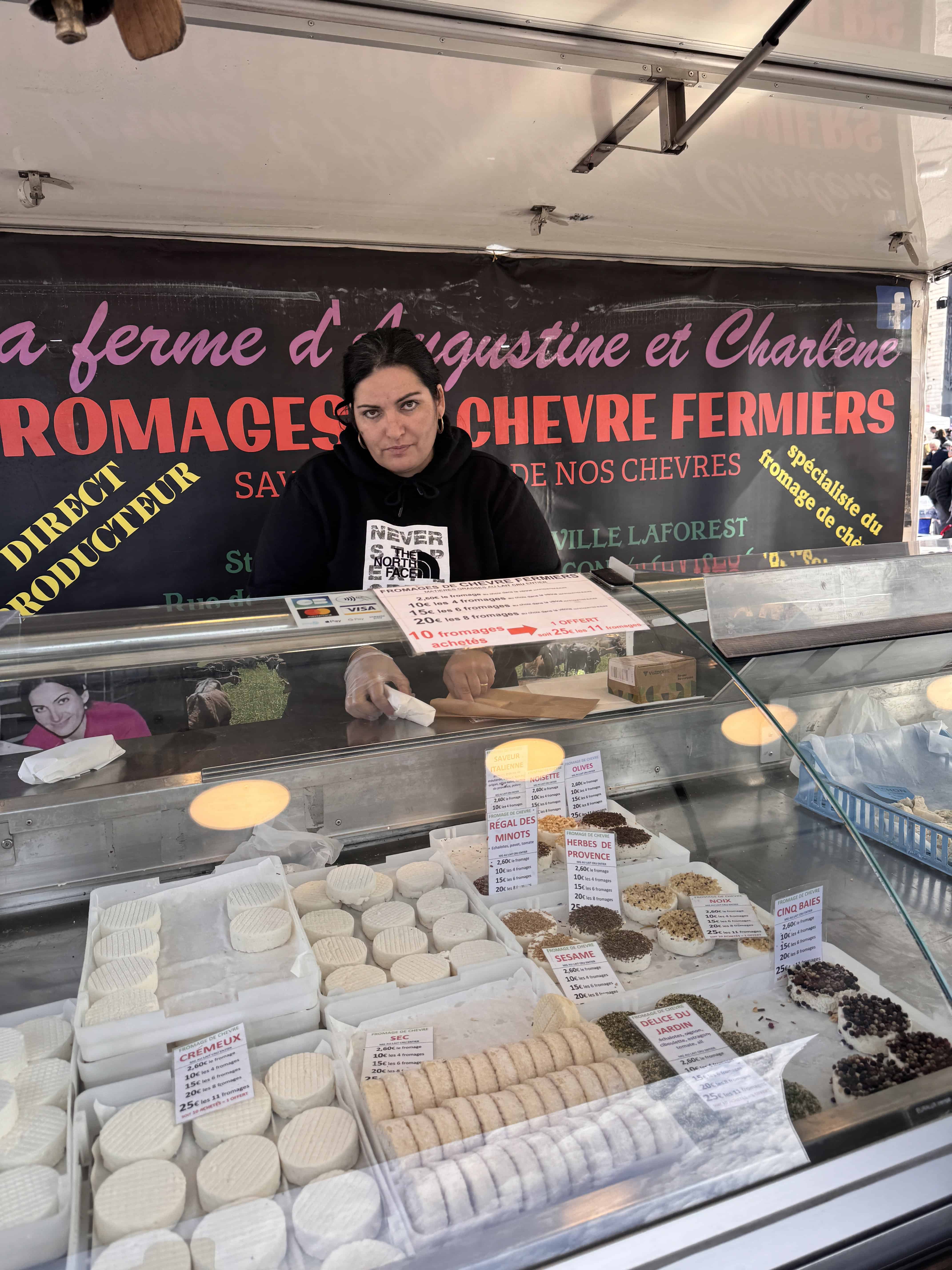 A woman sells various goat cheeses at an outdoor market stall in France, with a sign in French behind her—an authentic stop to add to your Marseille itinerary.