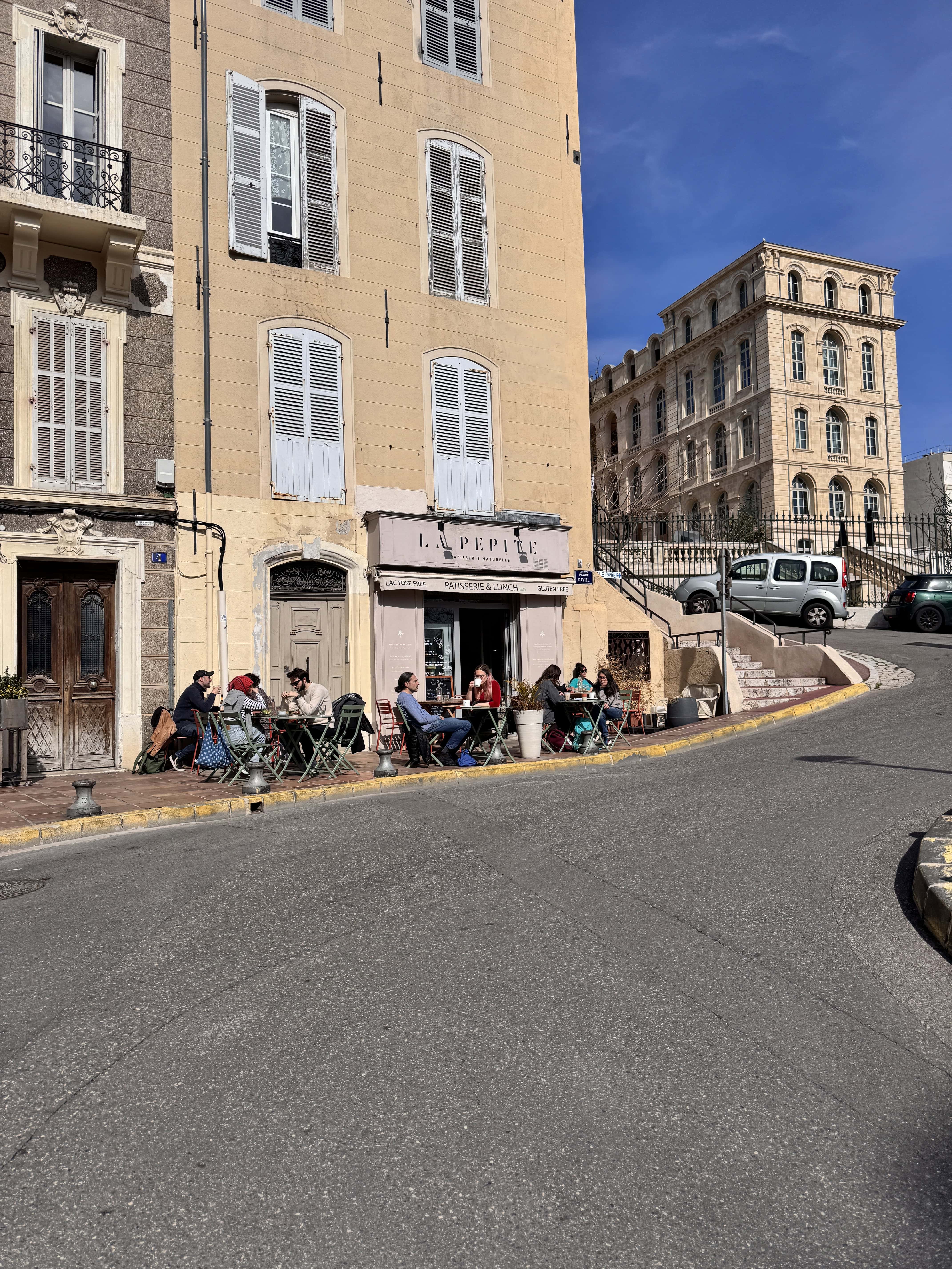 People sitting outside a café on a sunny day beside a sloped street with historic buildings in the background—a perfect Marseille scene to include in your France itinerary.