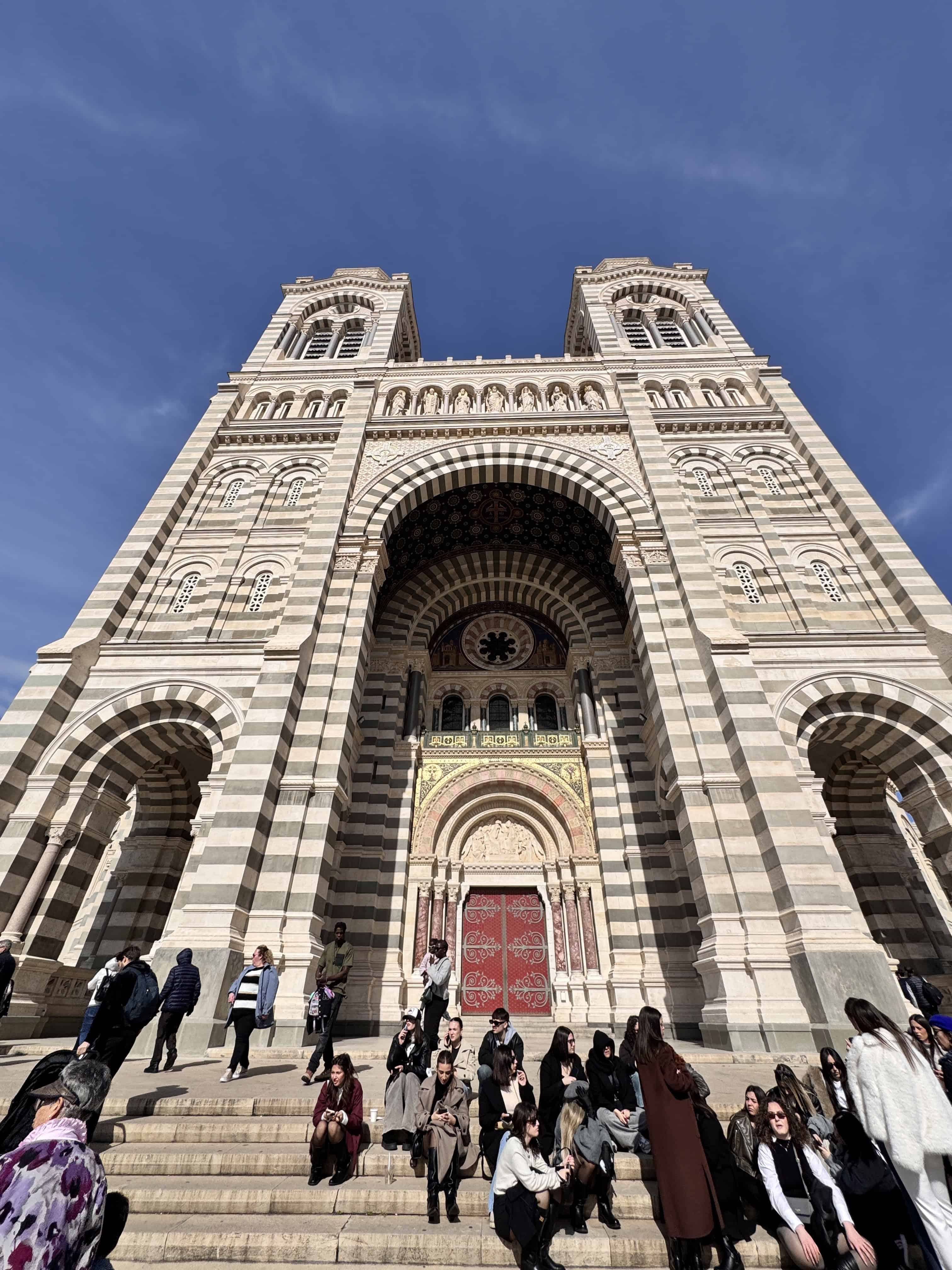 People sit and gather on the steps of a large striped stone cathedral under a blue sky in Marseille, France—a must-see stop on any itinerary.