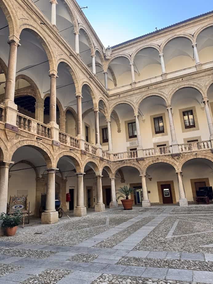Open courtyard with stone arches and columns, surrounded by a two-story building under a clear blue sky&mdash;a must-see when exploring what to do in Palermo for a day.