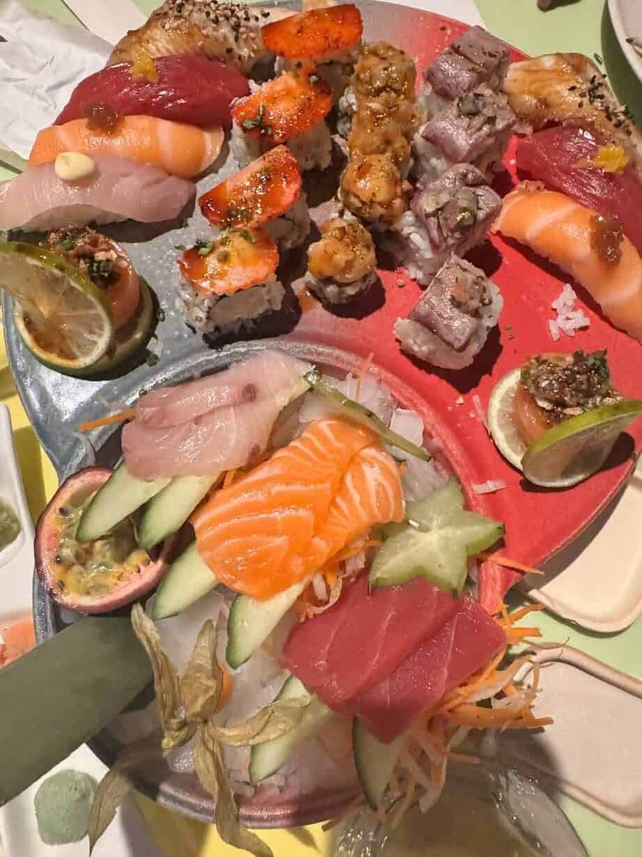 A colorful platter of assorted sushi and sashimi with garnishes, fruits, and vegetables on a red and gray plate, inspired by the vibrant fresh food found in Barcelona markets.
