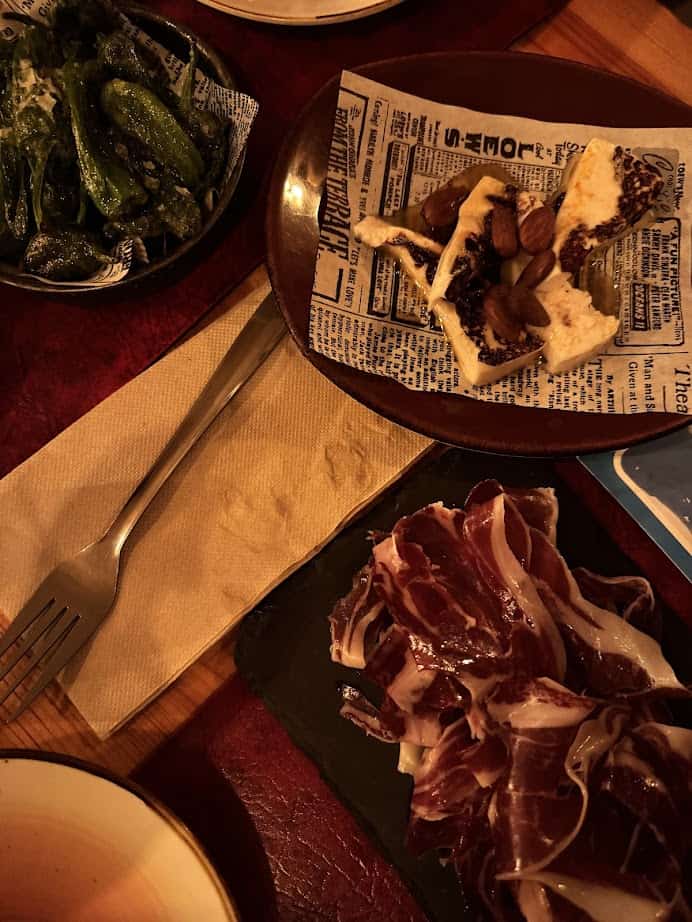 A table with plates of fresh food like green peppers, cheese with nuts, and slices of cured ham, plus a fork and napkin—perfect inspiration from Barcelona markets.