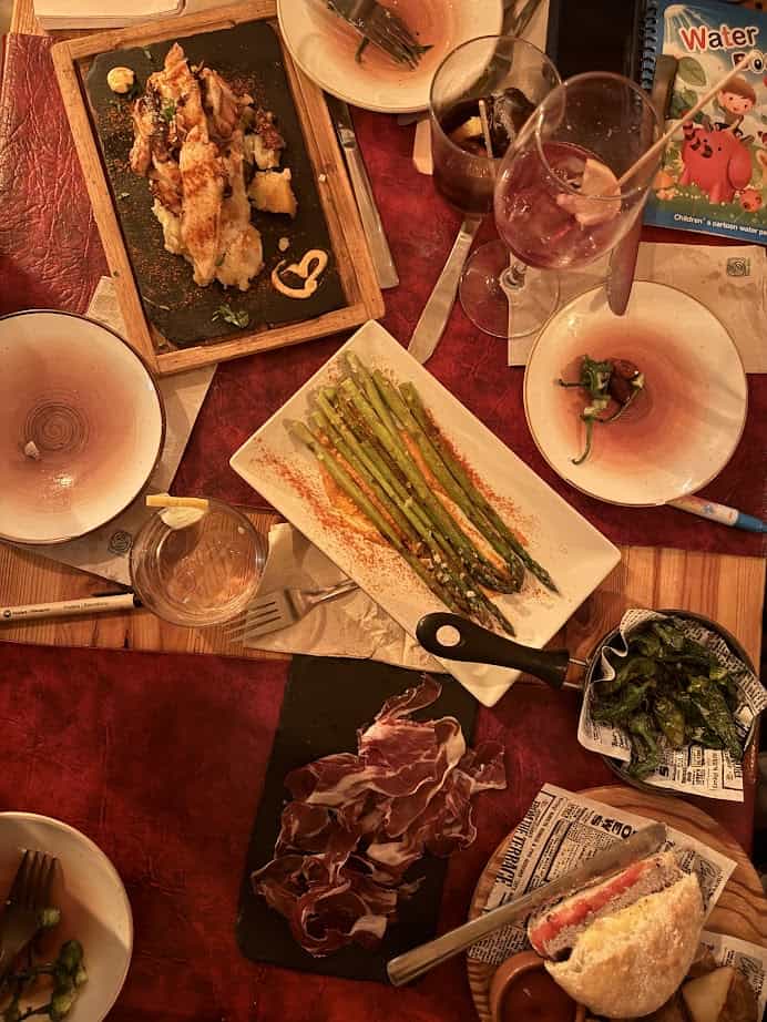 A table with fresh food—like asparagus, ham, bread, grilled seafood—and drinks is set on a red tablecloth, evoking the vibrant flavors found at Barcelona markets.