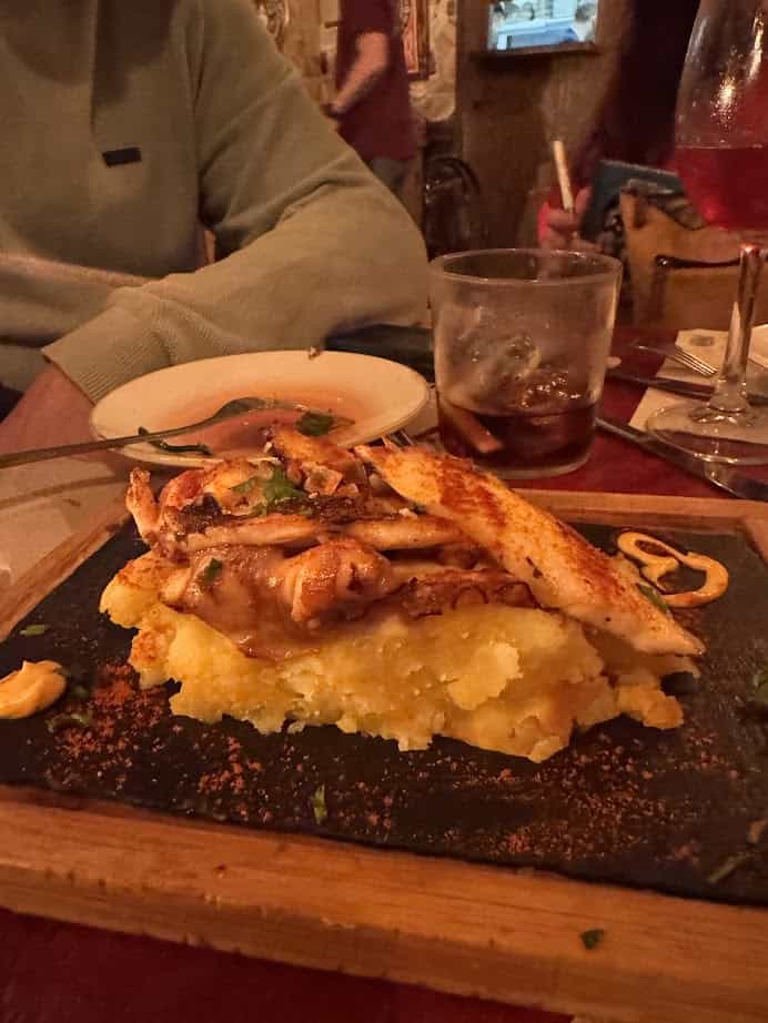 A wooden platter with grilled crab atop mashed potatoes, served on a restaurant table with drinks in the background—a delicious example of fresh food you might find at Barcelona markets.