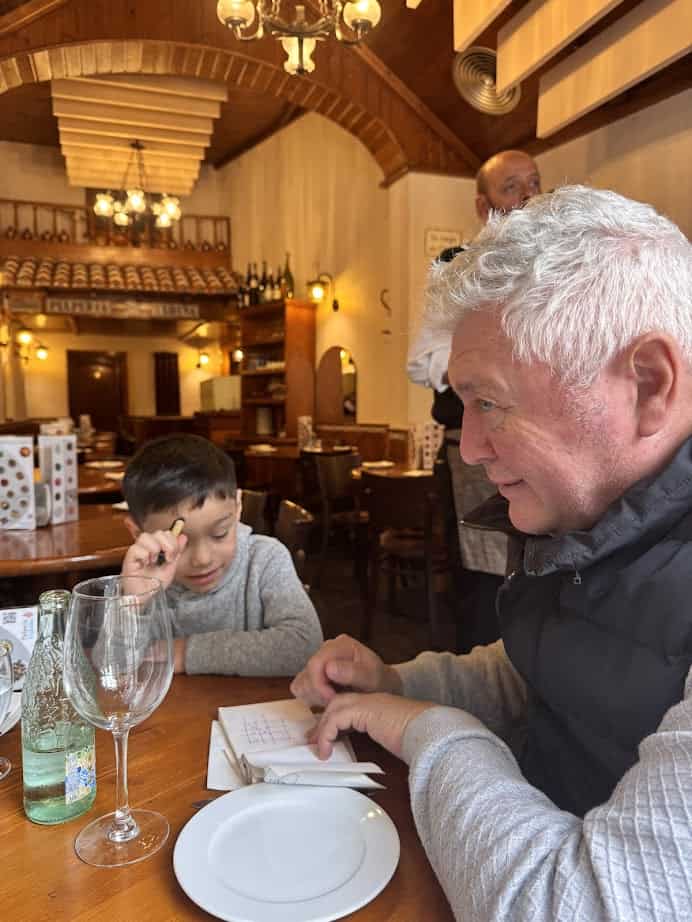 An older man and a boy play tic-tac-toe at a restaurant table, smiling as they enjoy their time together after exploring Barcelona markets and sampling fresh food.