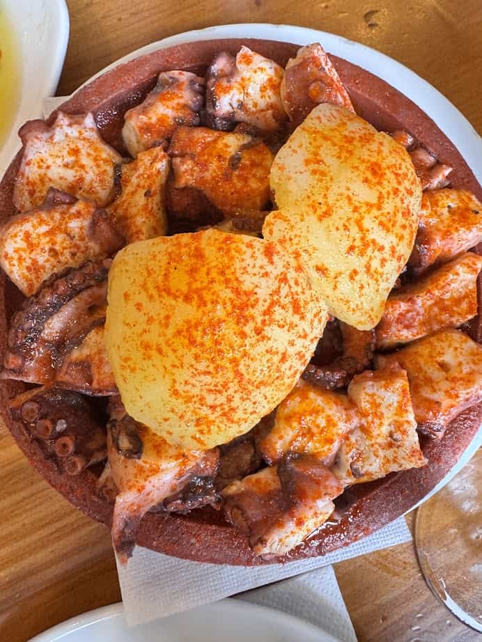 A plate of octopus topped with potatoes and sprinkled with paprika, inspired by the fresh food found in Barcelona markets, sits on a wooden table.