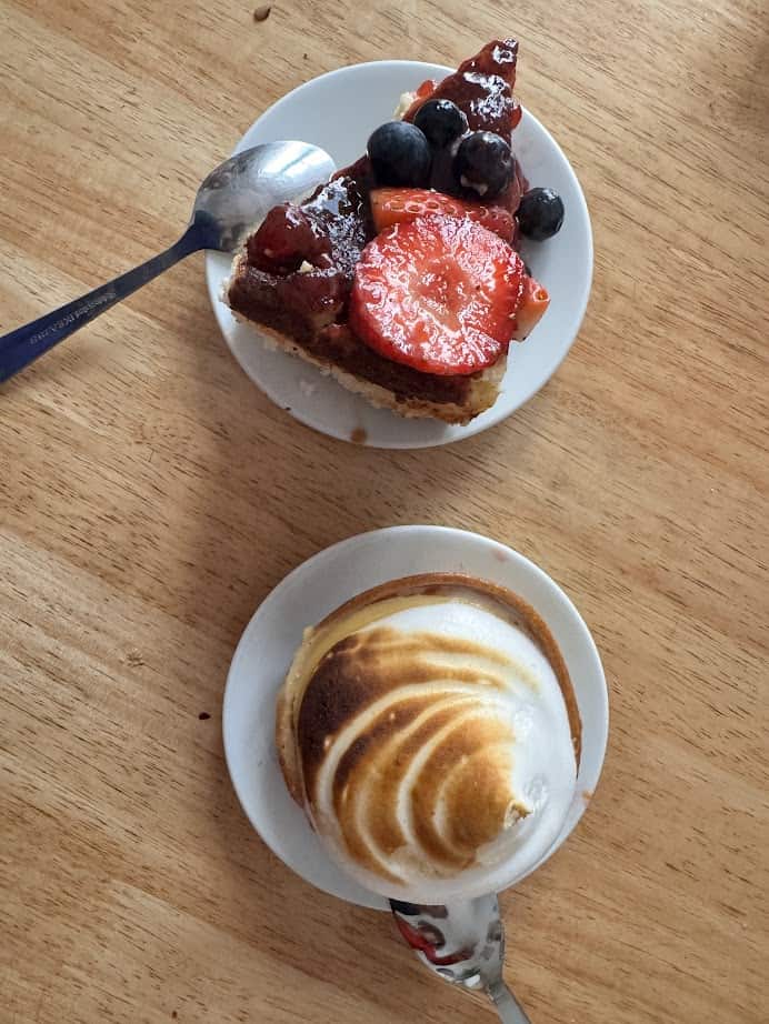 A slice of fruit tart and a lemon meringue tart on small plates with spoons on a wooden table, perfect for anyone seeking fresh food inspired by Barcelona markets.
