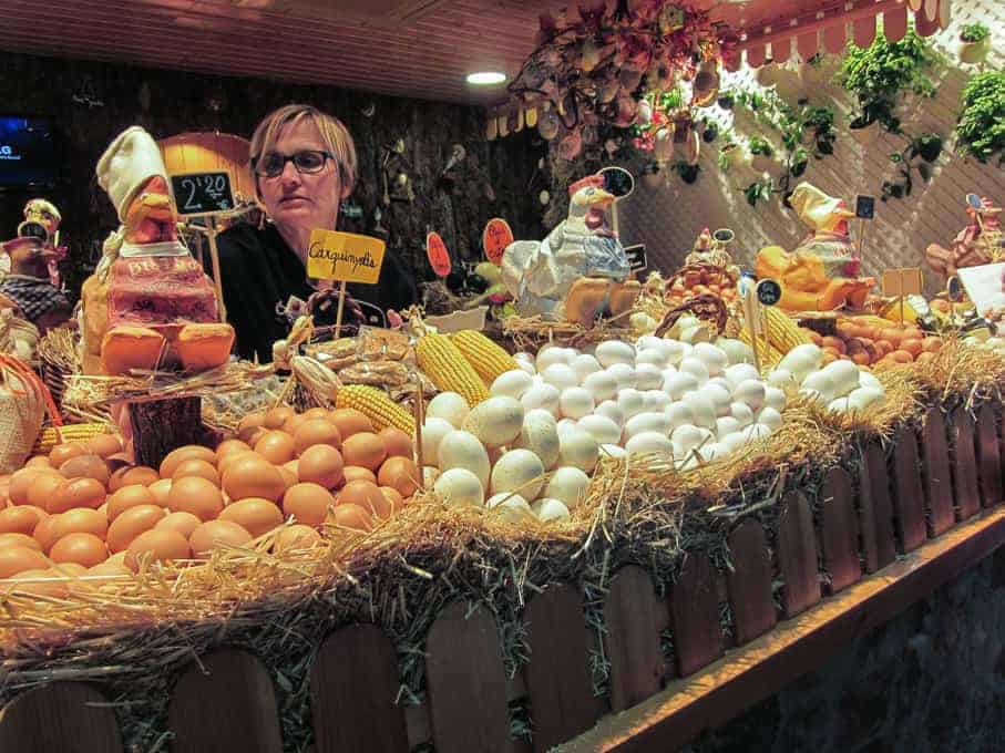 A woman stands behind a rustic market stall displaying eggs, corn, and various cheeses.