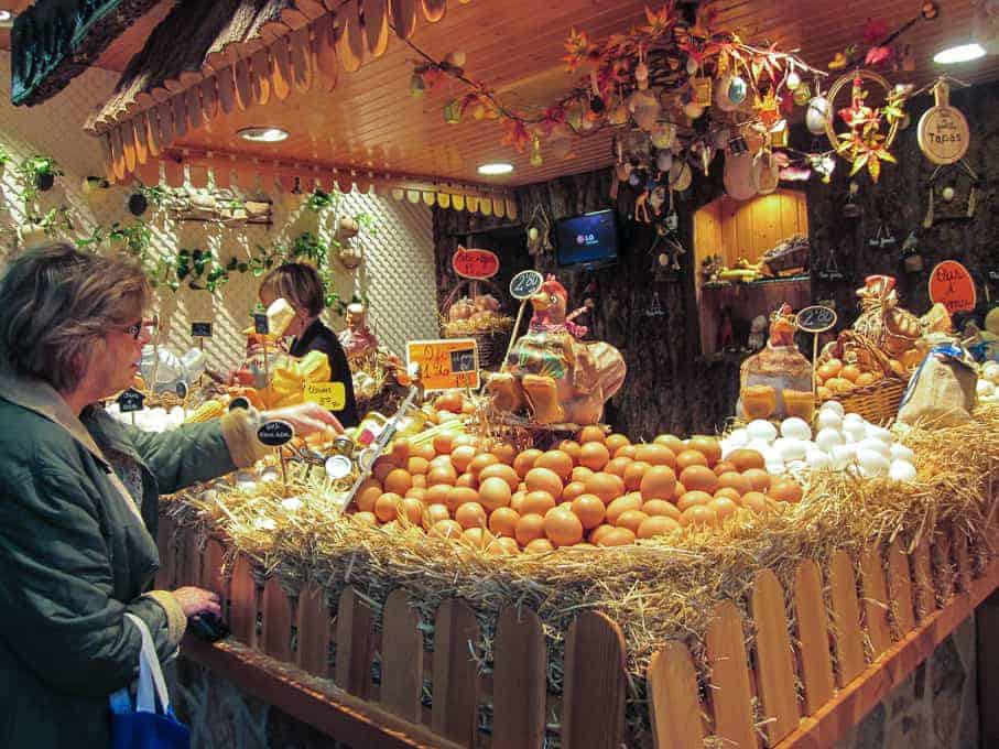 A woman selects eggs at a market stall displaying brown and white eggs on straw under warm lighting.
