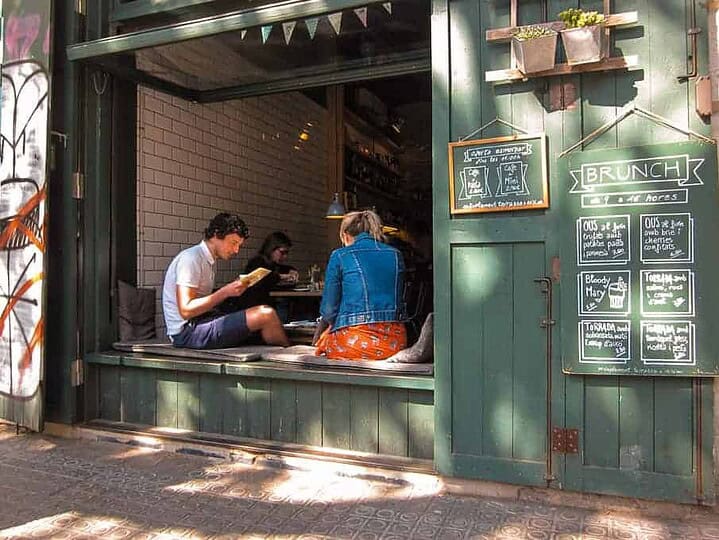 Two people sit by a large open window at a café, reading menus near bright green walls and chalkboard signs.