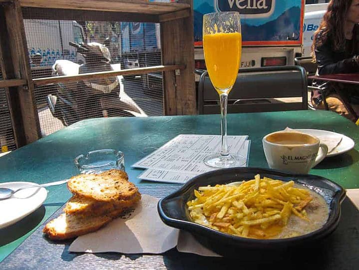Breakfast of toast, coffee, juice, and a dish topped with fries on a green outdoor table.