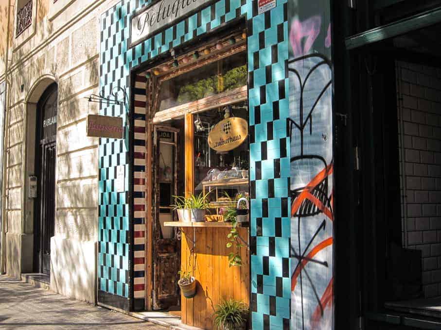Entrance of a barbershop with turquoise tiles and graffiti, plants on a wooden stand, and a glass window.