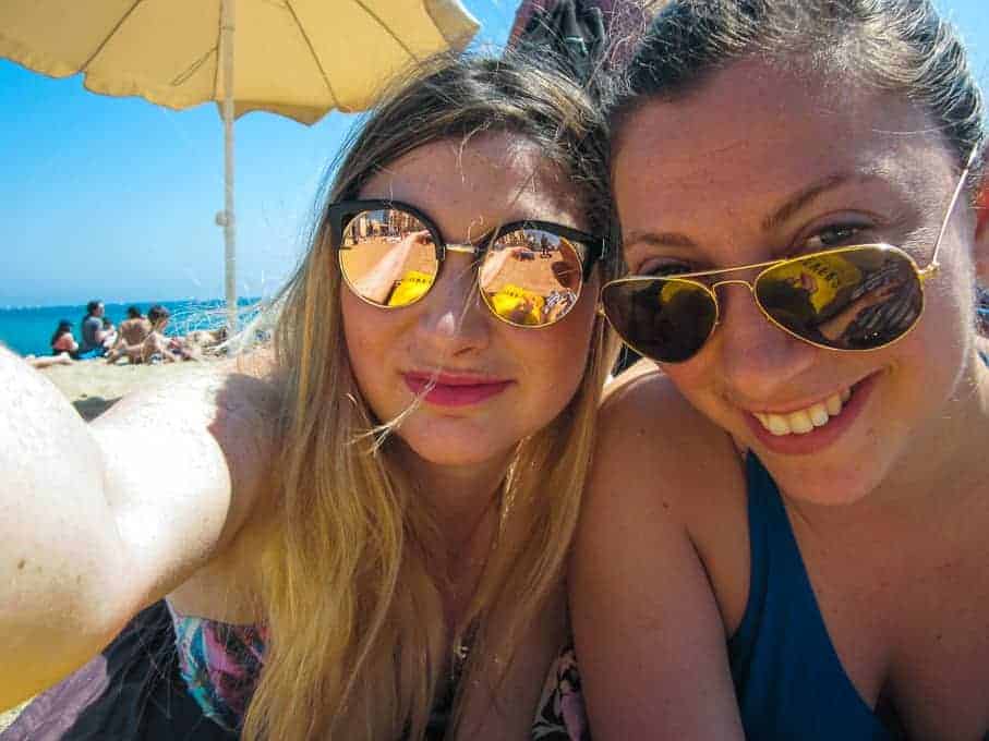 Two women in sunglasses smiling and taking a selfie on a sunny beach under a large umbrella.