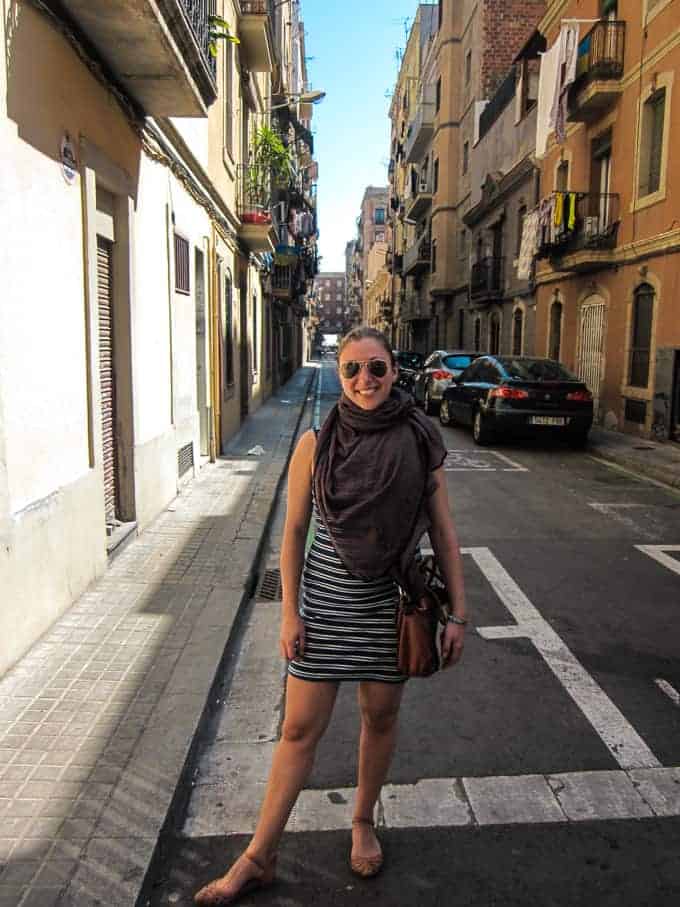 Woman in a striped dress stands smiling on a sunny, narrow street lined with buildings and parked cars.