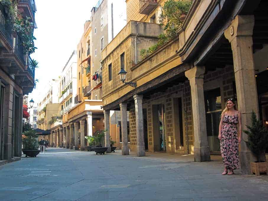 A woman in a floral dress stands on a quiet, sunlit street lined with old buildings and columns.