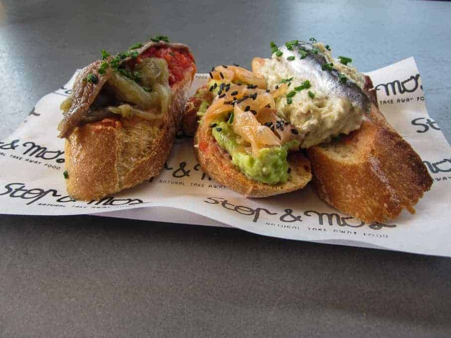 Three pieces of bruschetta topped with various spreads, vegetables, and fish on branded paper.