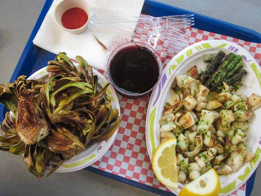 Grilled artichokes, roasted potatoes with asparagus, lemon wedges, red sauce, and a drink on a tray.