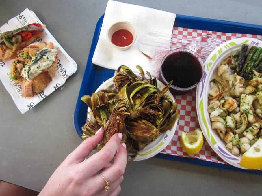 A hand grabs fried artichokes on a plate next to wine, grilled veggies, and a crostini on a table.