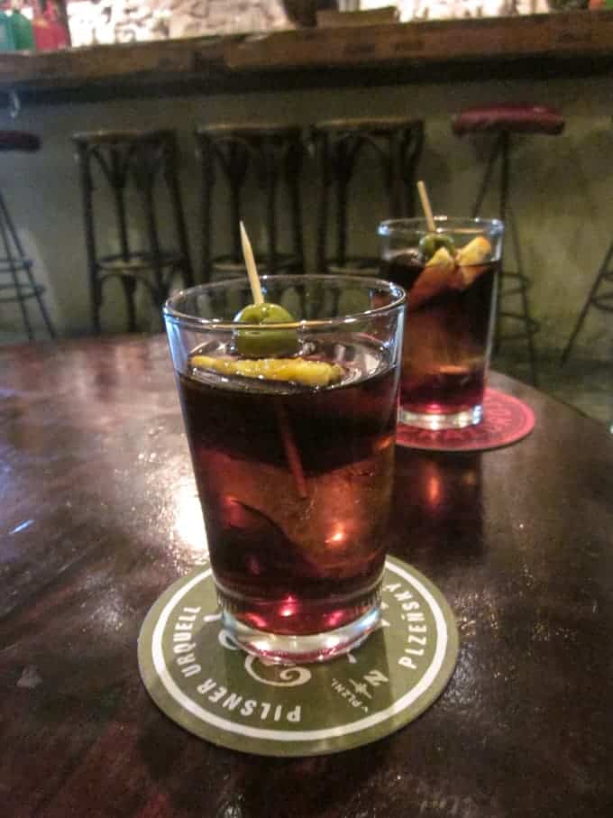 Two glasses of dark cocktail with olives and orange slices on coasters, on a wooden table in a bar.