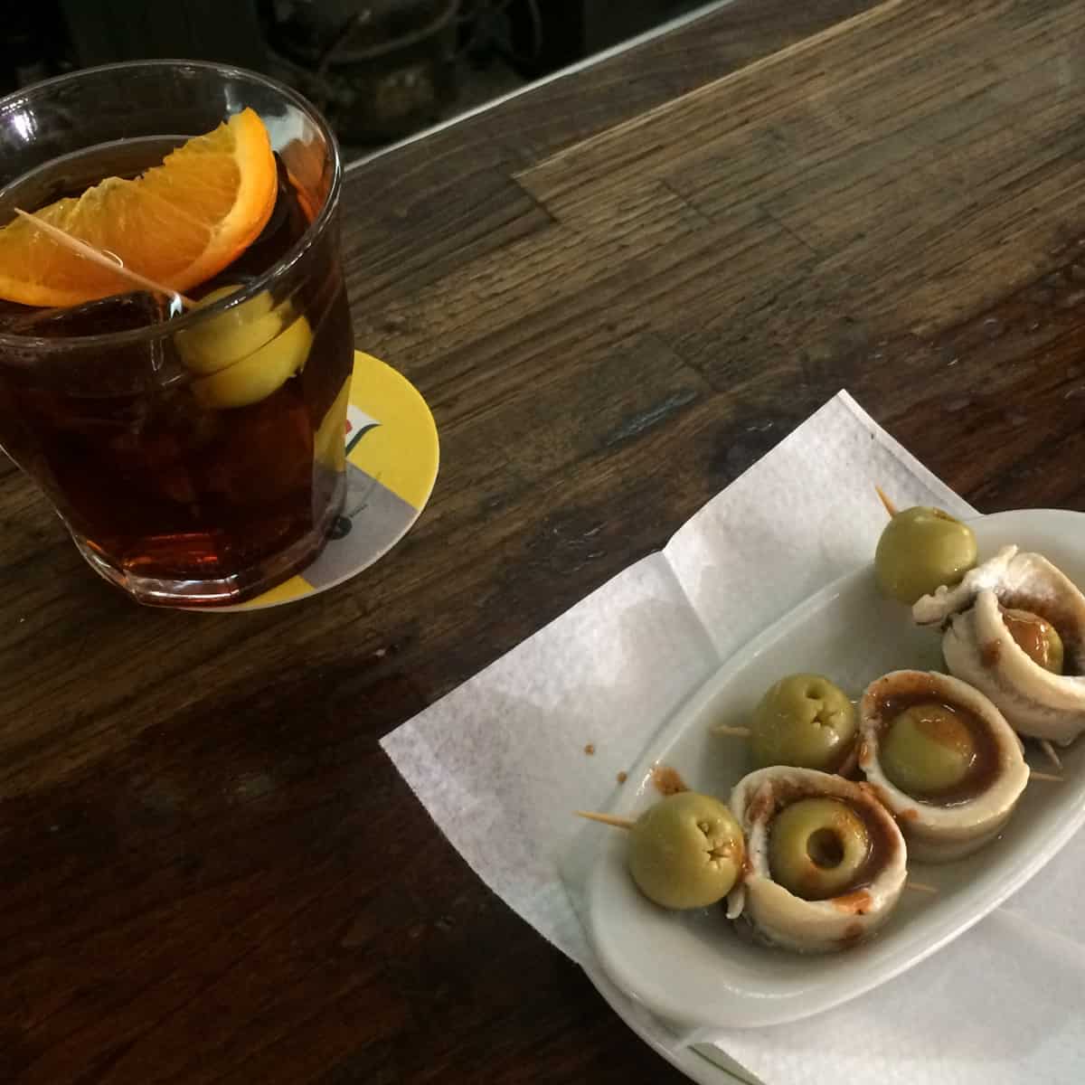 Glass of dark drink with an orange slice, and a plate of olives wrapped in anchovies on a napkin.