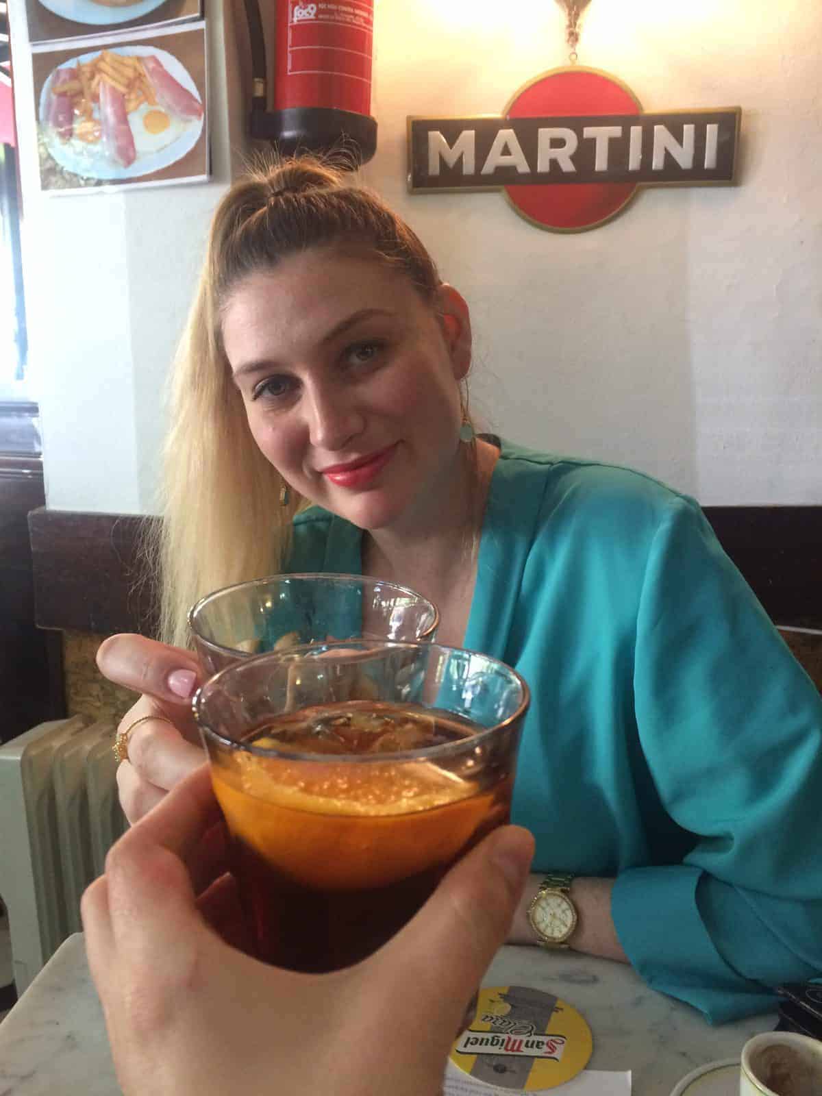 A woman in blue smiles while toasting glasses with another person at a table in a café.