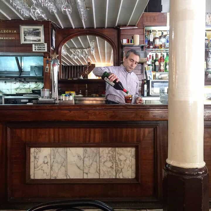 A bartender pours a drink behind a wooden bar in a classic, vintage-style bar or café.