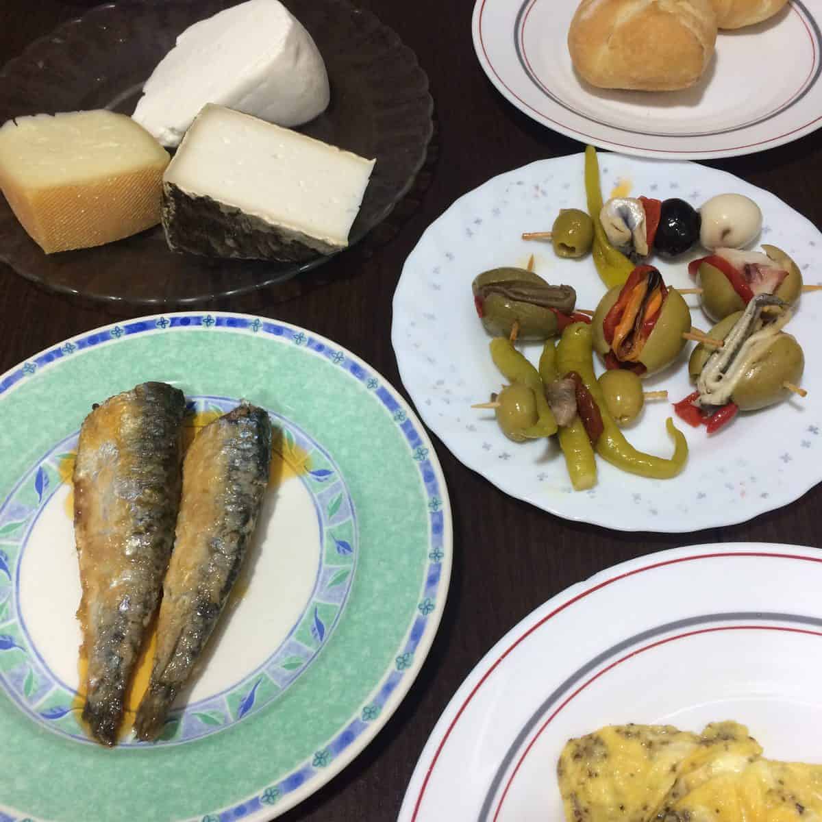 Plates of cheese, bread rolls, sardines, marinated vegetables, and an omelet on a dark table.