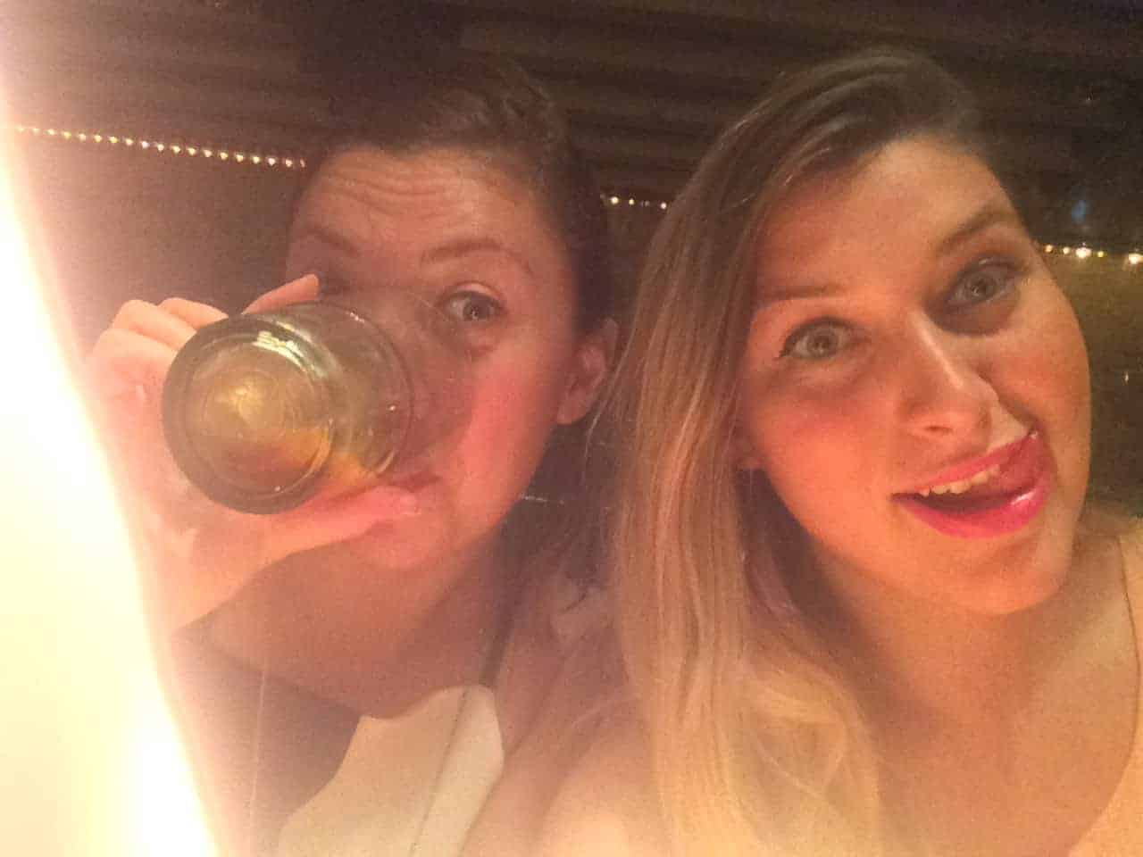 Two women pose for a selfie; one is drinking from a glass, and the other is smiling playfully.