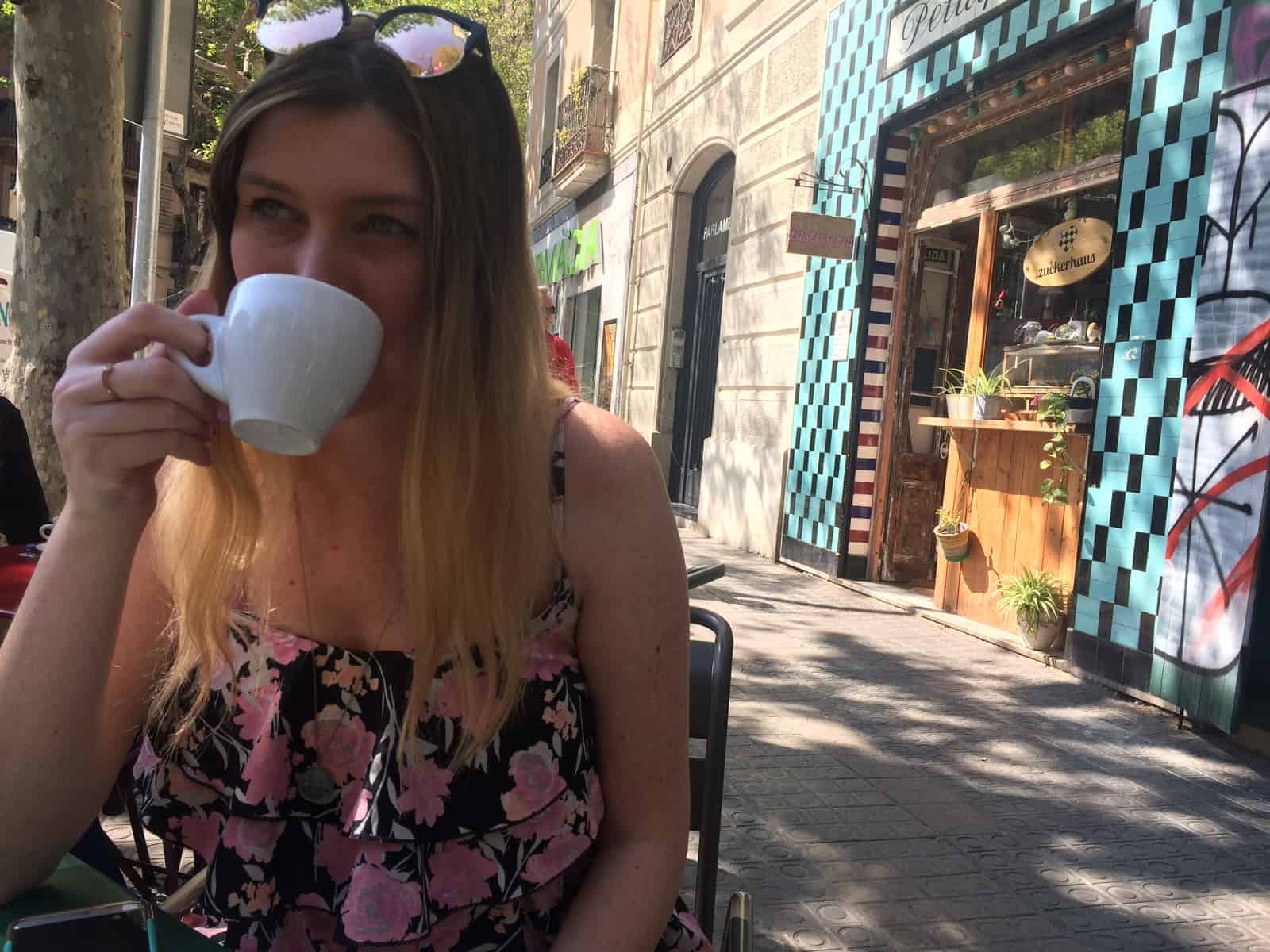 Woman in a floral dress drinks coffee at an outdoor café with colorful buildings in the background.