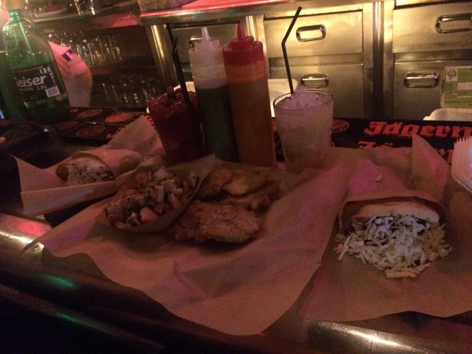 Dimly lit bar counter with sandwiches, fries, salad, condiments, and drinks arranged on parchment paper.