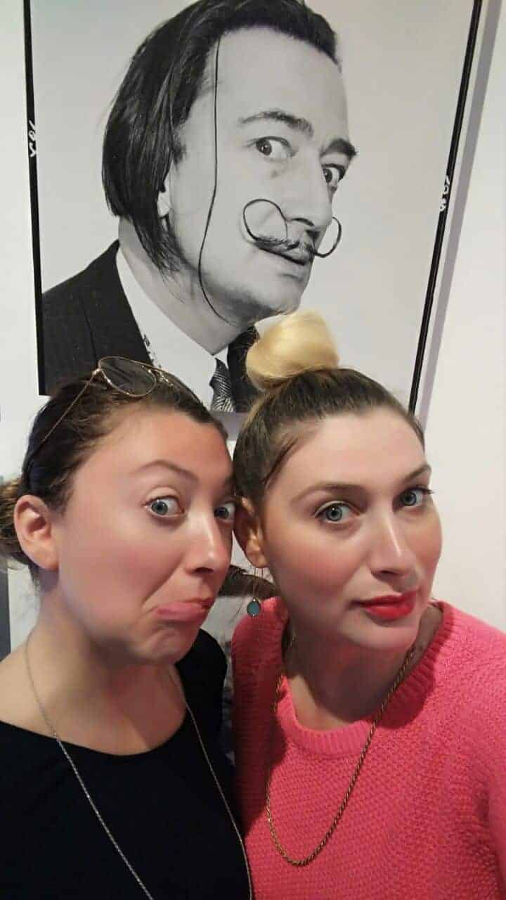 Two women pose, mimicking faces, in front of a black-and-white Salvador Dalí portrait.