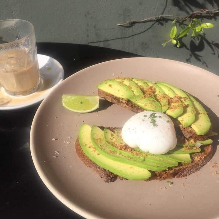 Avocado toast with a poached egg and lime on a plate, next to a glass of coffee on a saucer.
