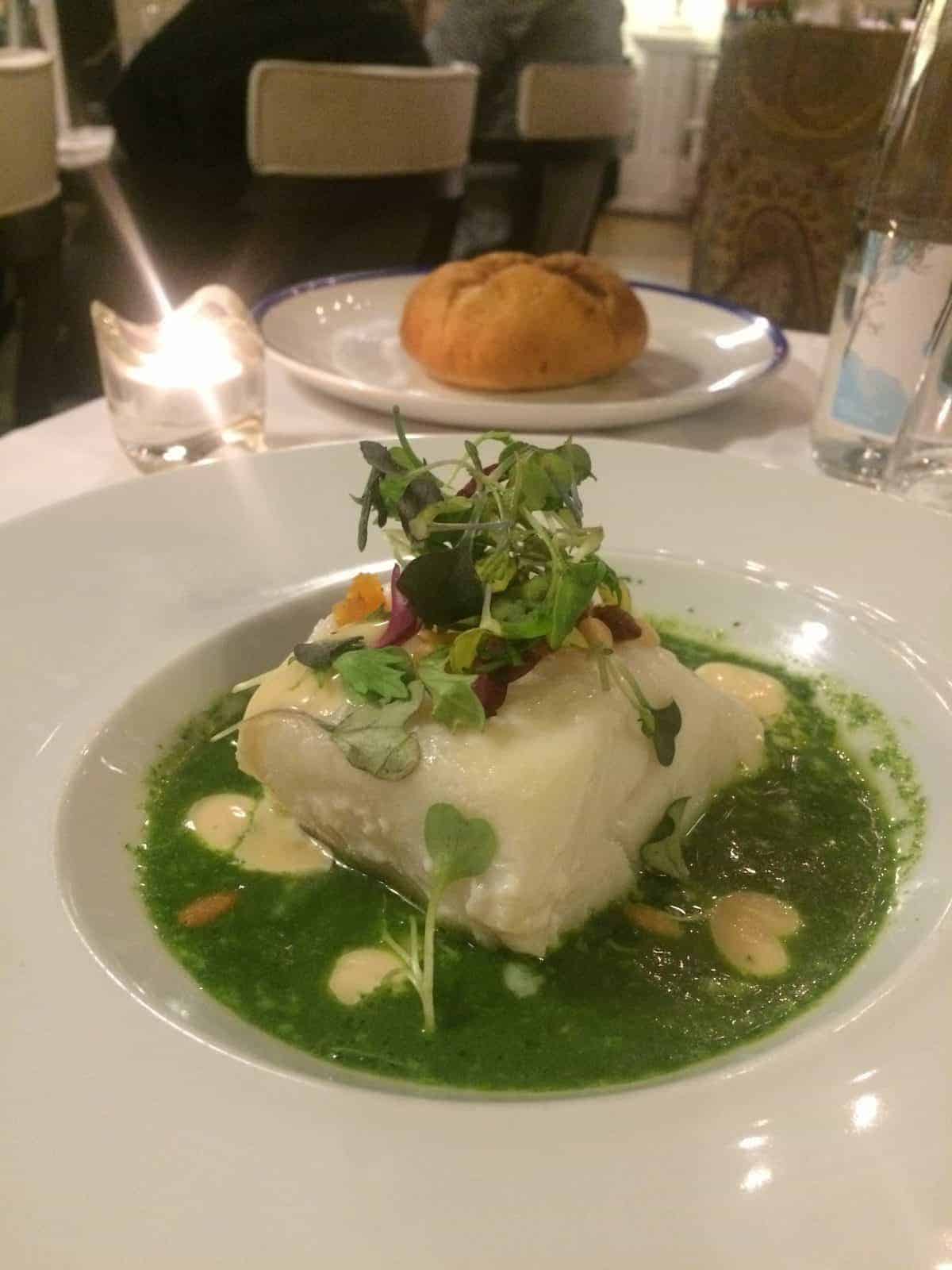 A plated fish fillet with green sauce and microgreens, with bread and a lit candle in the background.