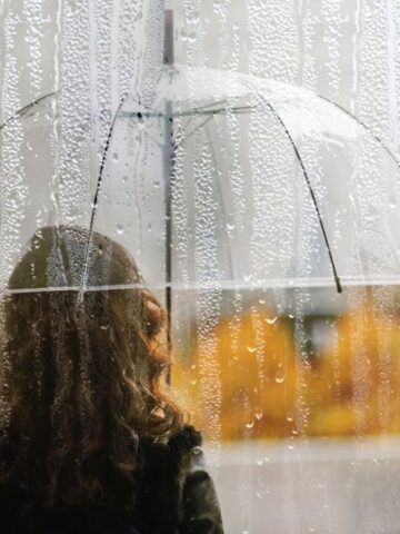 A person with an umbrella stands in front of a rain-covered window, with autumn colors in the background.