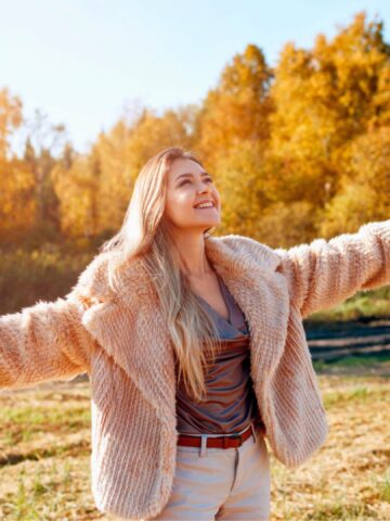 Smiling woman in a fur coat stands outdoors with arms outstretched, surrounded by autumn trees.