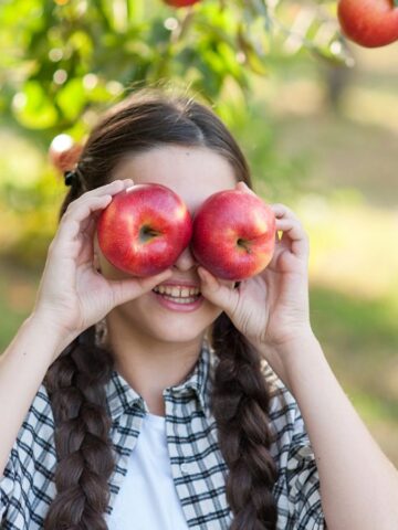 Girl with braids holding two apples over her eyes, smiling in an orchard with apples on the tree.